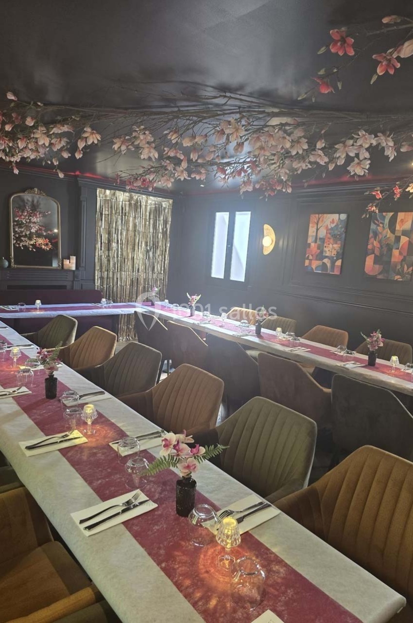 Salle de réception élégante avec tables dressées, nappes blanches et roses, décor floral suspendu et lumière tamisée.