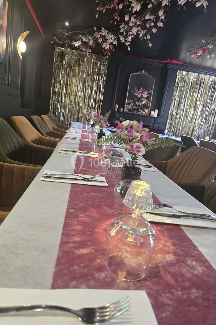 Table dressée avec nappe rose, verres, couverts et décor floral dans une salle élégante aux tons sombres et dorés.