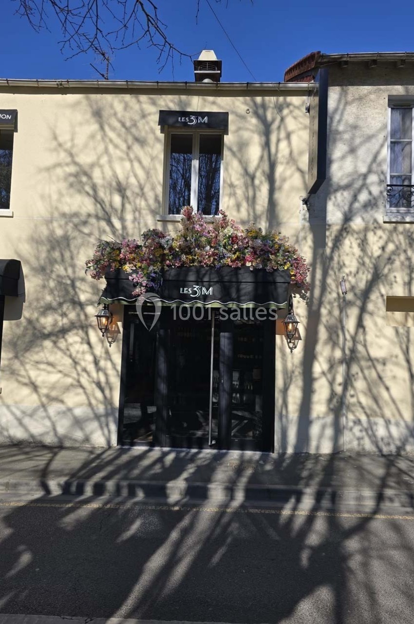 Façade d'un bâtiment beige avec une enseigne ’Les 3M’, ornée de fleurs colorées au-dessus de l'entrée.