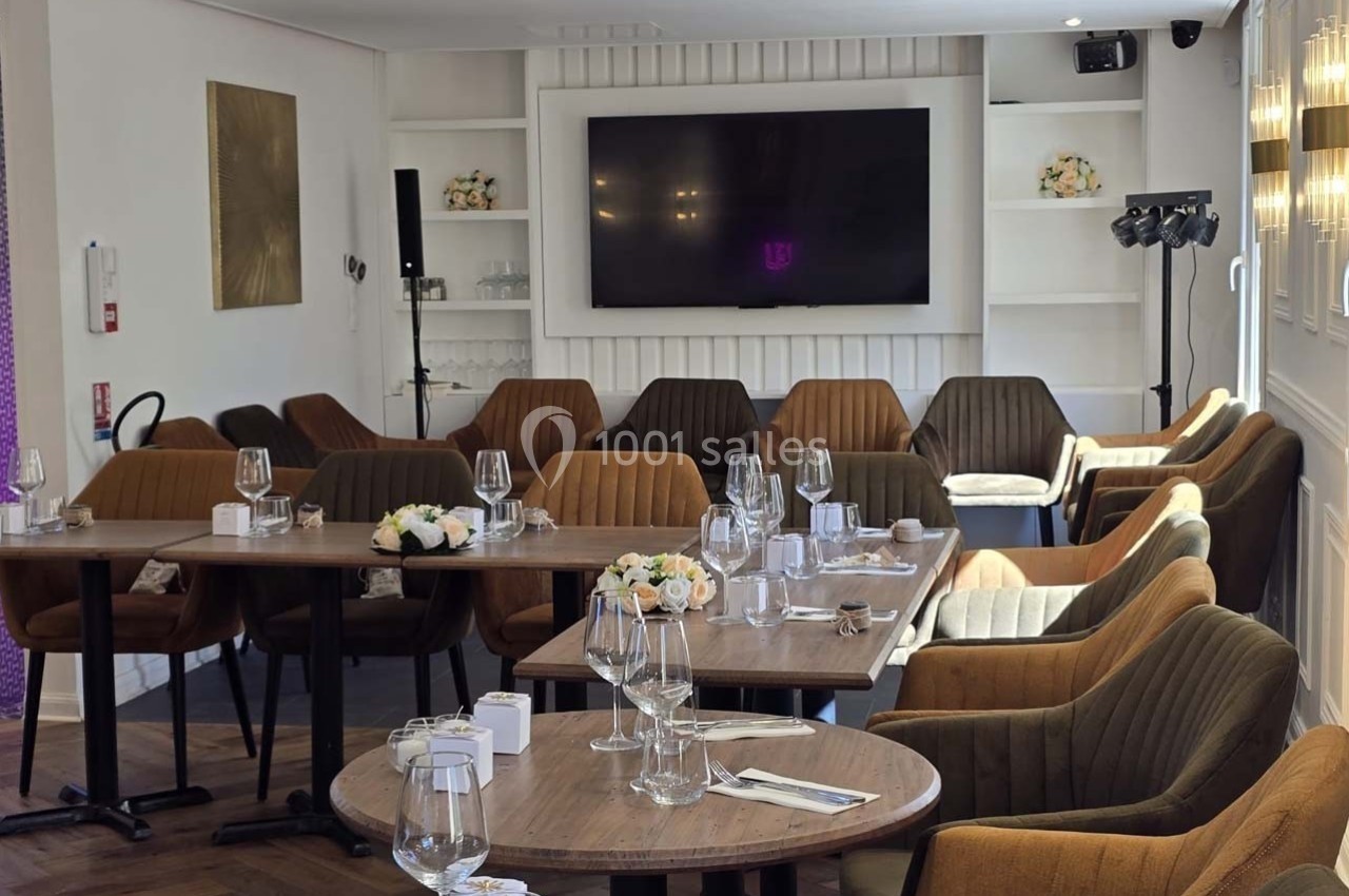 Salle de restaurant lumineuse avec tables dressées, chaises confortables et écran mural en arrière-plan.