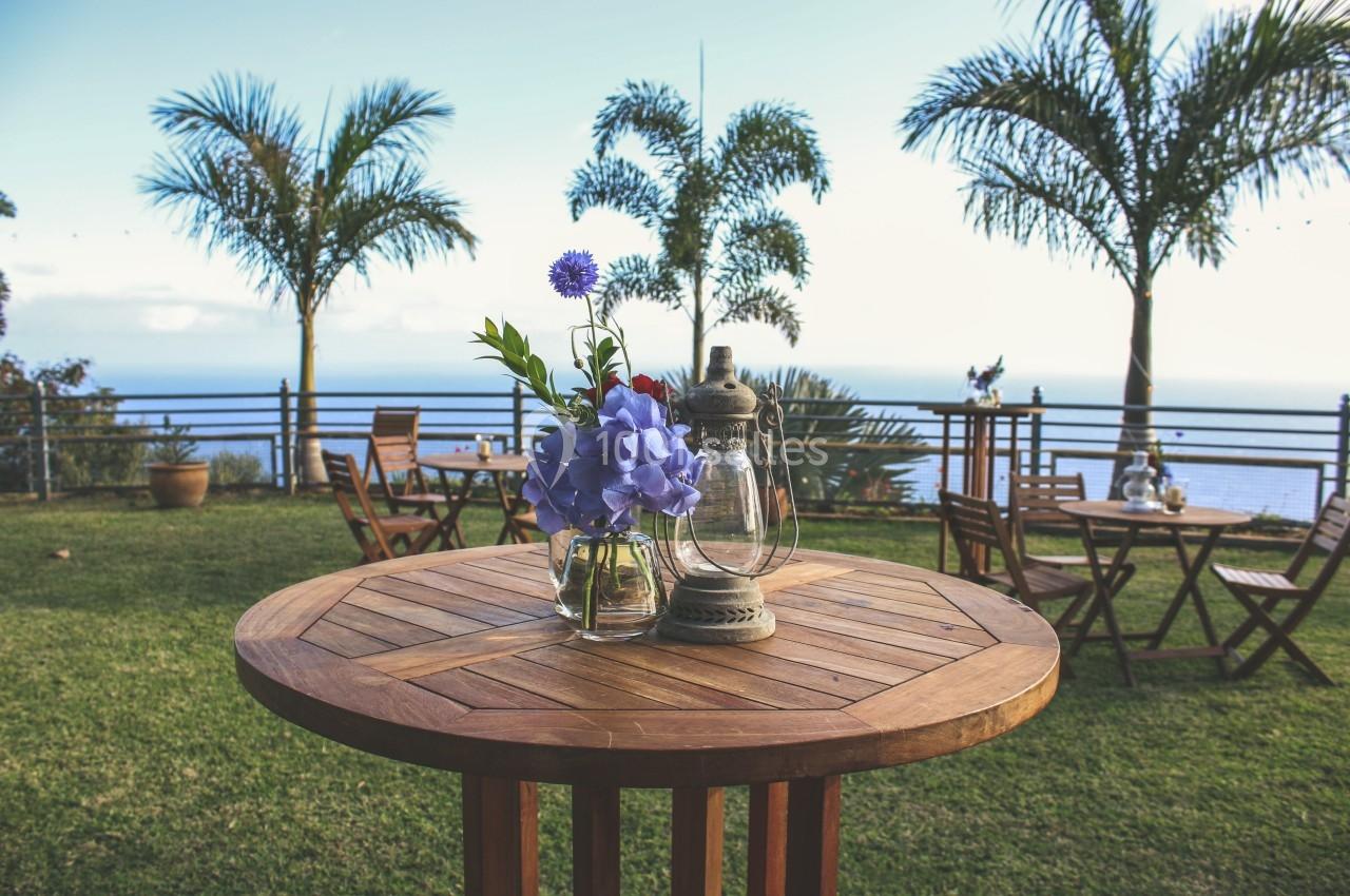 Table en bois avec un vase de fleurs et une lanterne, située dans un jardin avec vue sur la mer et des palmiers.