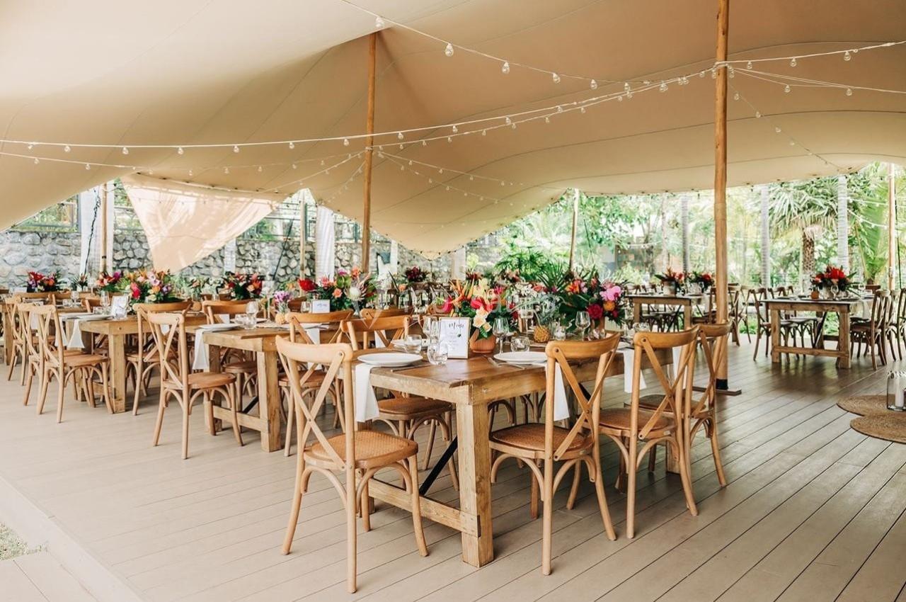Salle de réception en plein air avec tables en bois décorées de fleurs colorées sous une tente beige.