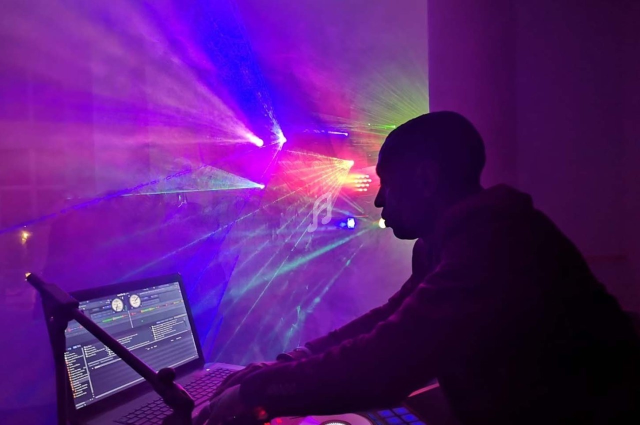 Un DJ mixe sur une console devant un ordinateur, entouré de faisceaux lumineux colorés dans une ambiance sombre.