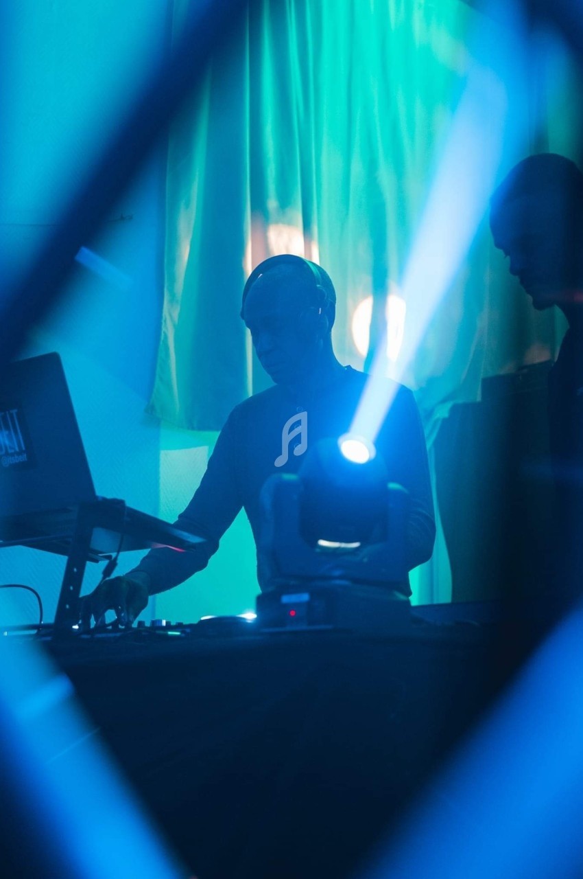 Deux DJ mixent sur une scène éclairée par des lumières bleues et un projecteur dans une ambiance nocturne.