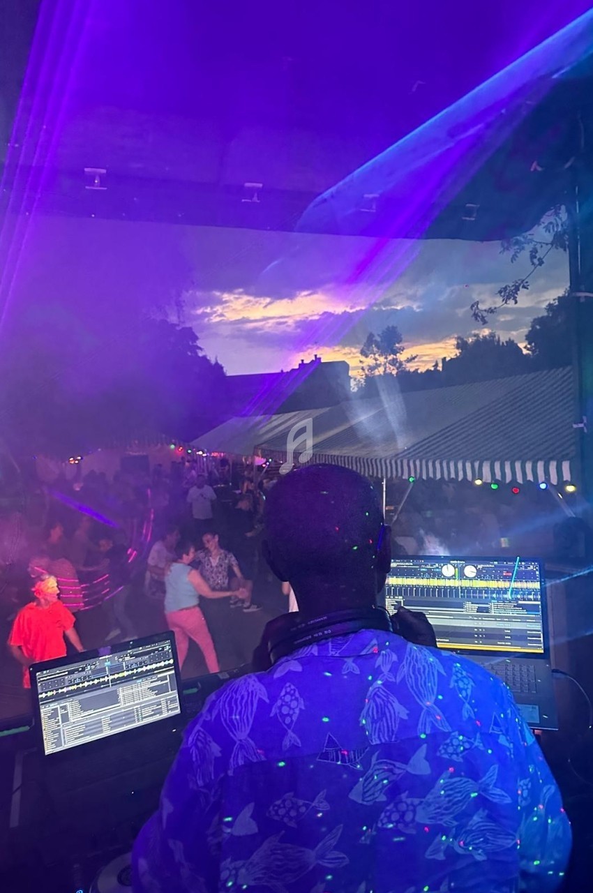 Un DJ mixe en plein air au coucher du soleil, face à une foule dansante sous des lumières colorées.