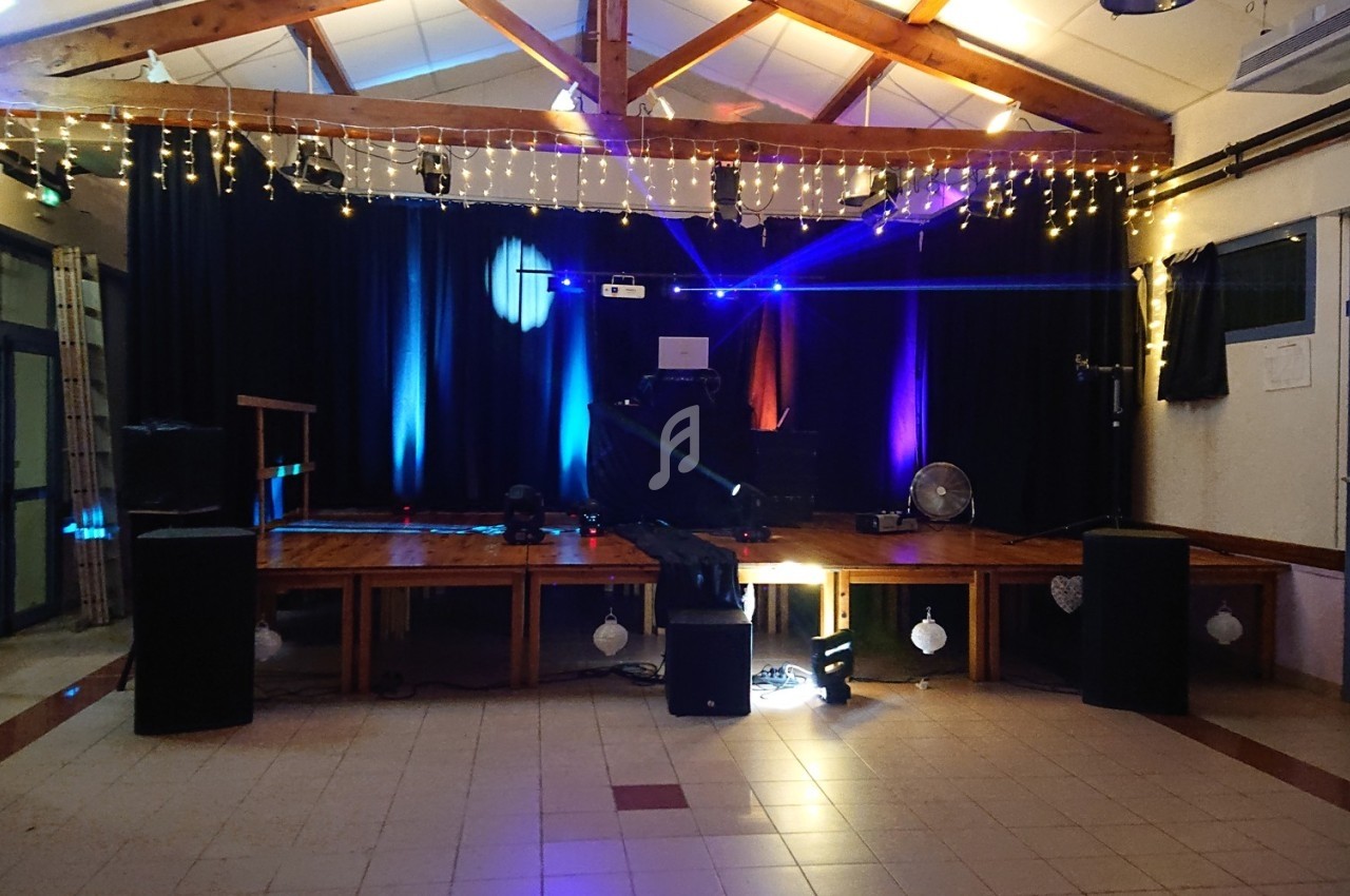 Scène décorée avec des guirlandes lumineuses, éclairages colorés et équipements audio dans une salle vide.