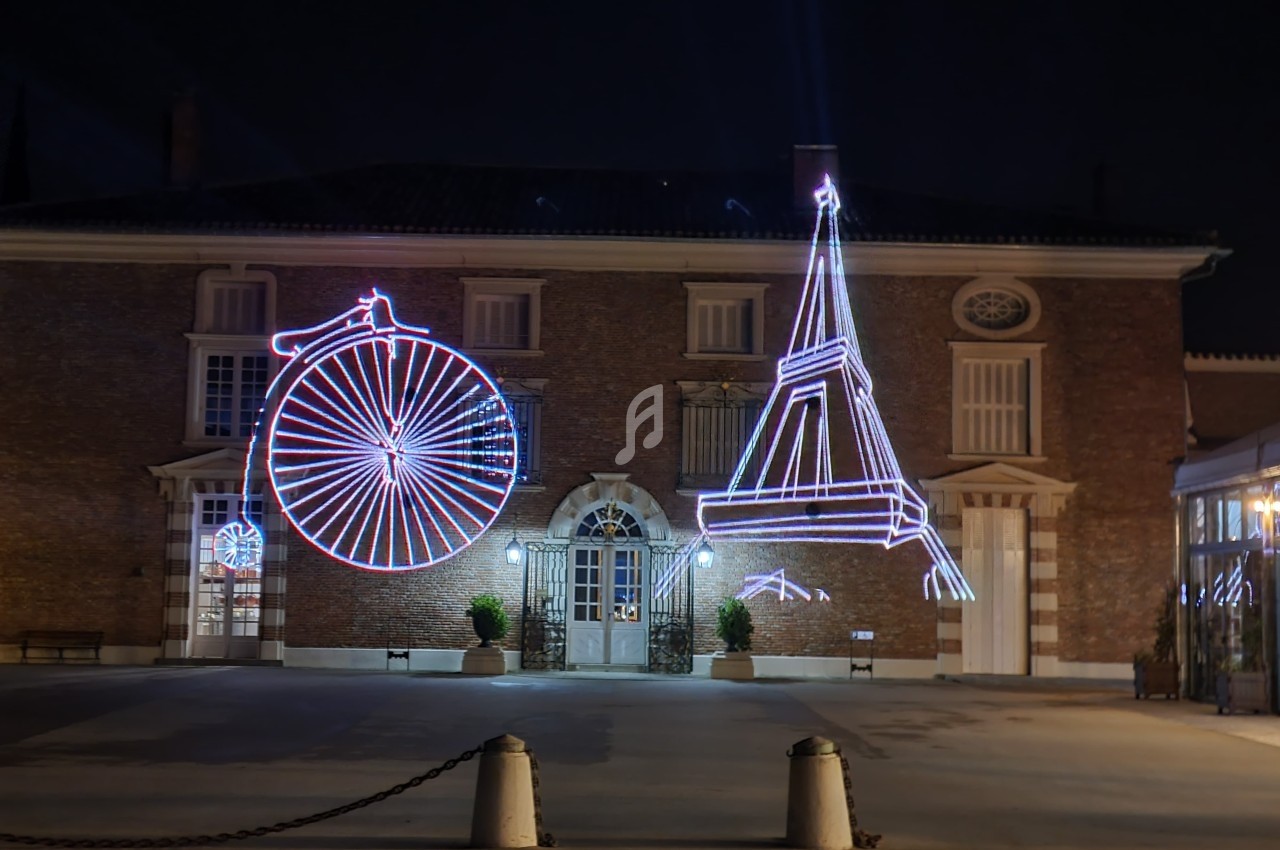 Façade d'un bâtiment en briques éclairée par des décorations lumineuses représentant un grand-bi et la tour Eiffel.