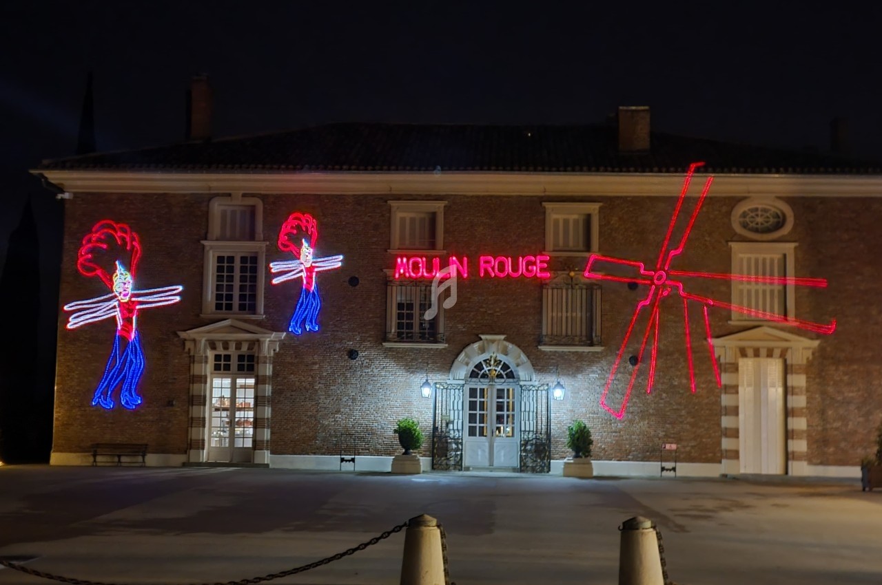 Façade d'un bâtiment en briques éclairée par des néons rouges représentant des danseuses et des ailes de moulin.