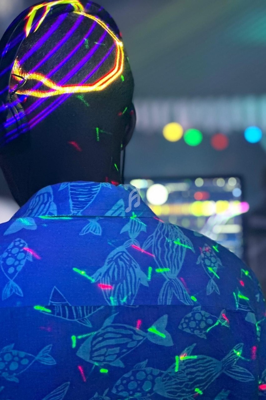 Homme de dos portant une chemise à motifs de poissons, éclairé par des lumières colorées dans une ambiance festive.
