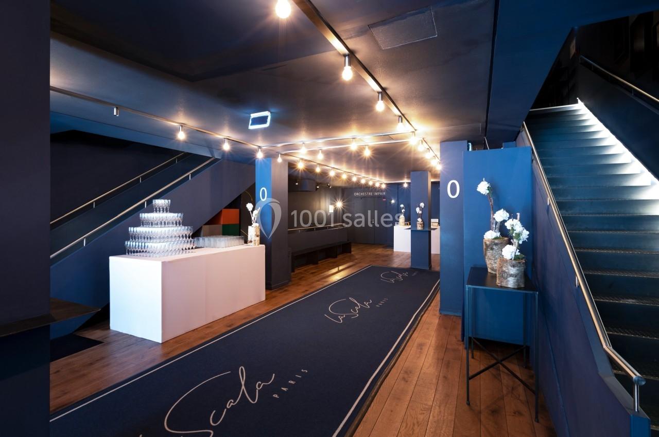 Hall d'entrée moderne avec éclairage suspendu, tapis bleu marqué ’La Scala’, escalier et tables décorées.