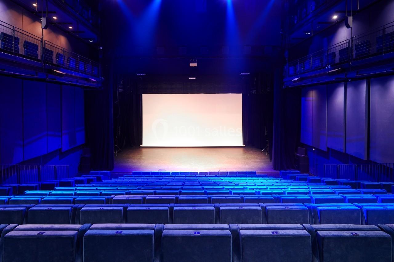Salle de théâtre vide avec des rangées de sièges bleus et une scène éclairée par des projecteurs.