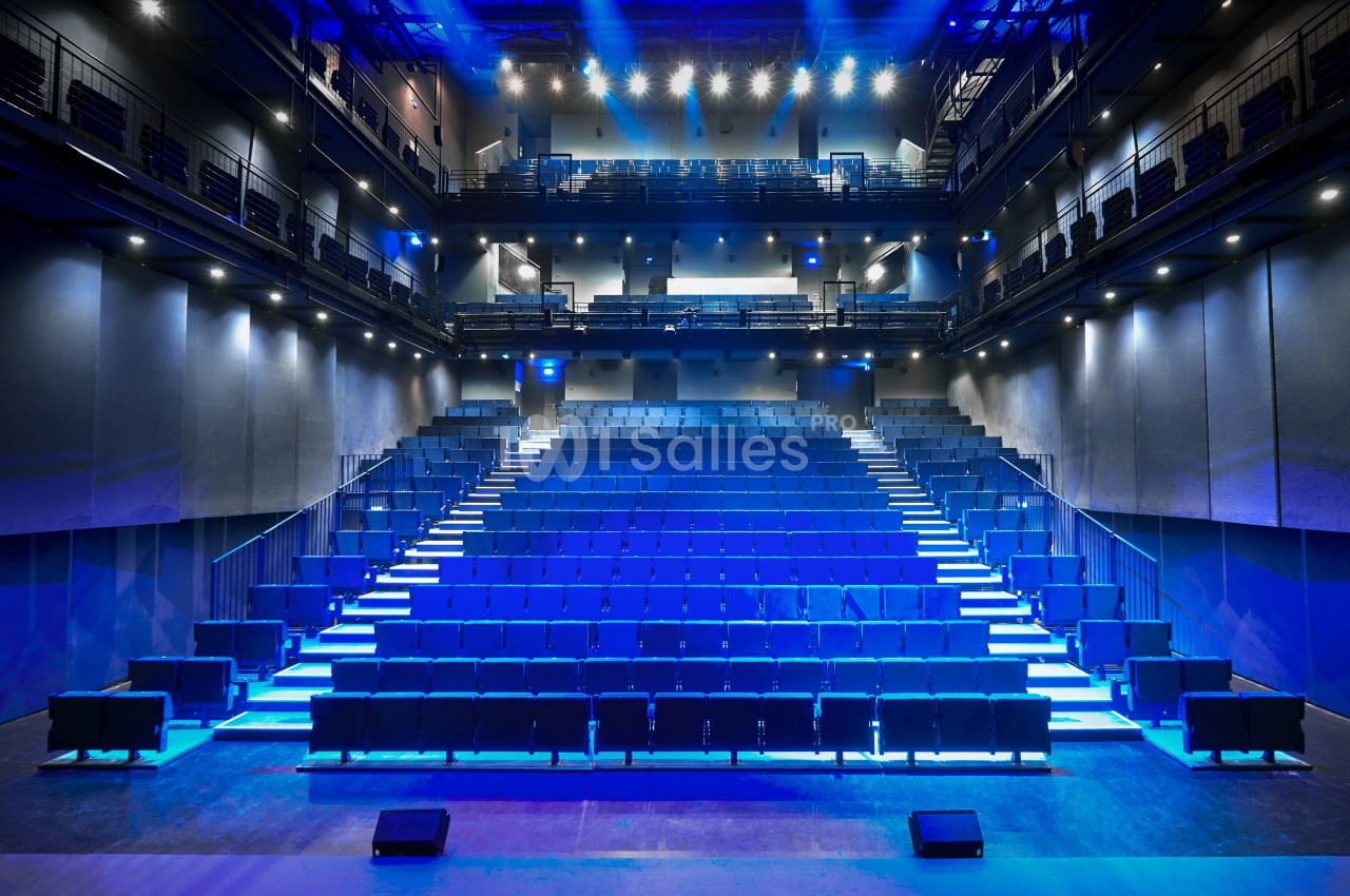 Salle de spectacle moderne avec gradins vides, éclairée par des lumières bleues et des projecteurs au plafond.