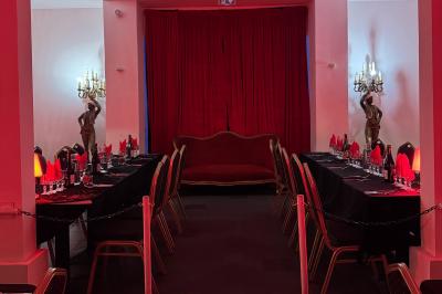 Salle décorée avec des rideaux rouges, tables dressées pour un repas, chaises noires et fauteuil doré.