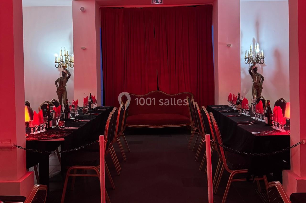 Salle décorée en rouge avec un canapé central, deux tables dressées et des statues éclairées par des appliques murales.