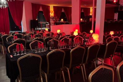 Salle décorée avec des rideaux rouges, tables dressées pour un repas, chaises noires et fauteuil doré.