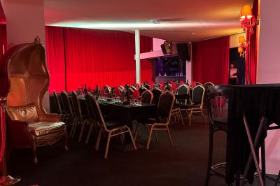 Salle décorée avec des rideaux rouges, tables dressées pour un repas, chaises noires et fauteuil doré.