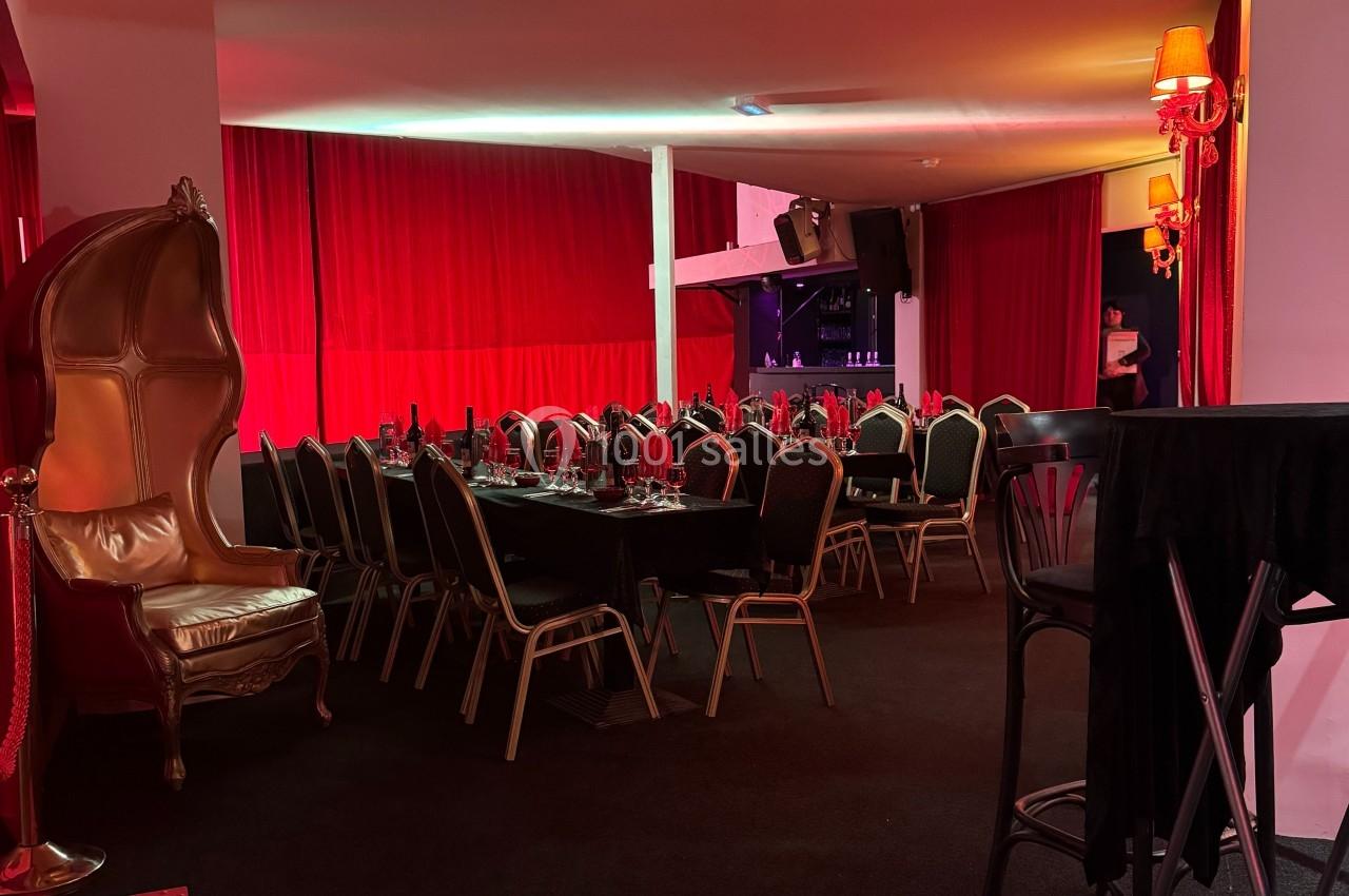 Salle décorée avec des rideaux rouges, tables dressées pour un repas, chaises noires et fauteuil doré.