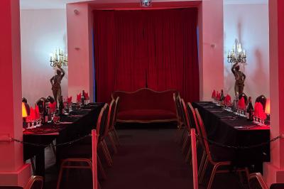 Salle décorée avec des rideaux rouges, tables dressées avec chaises noires et dorées, ambiance tamisée.