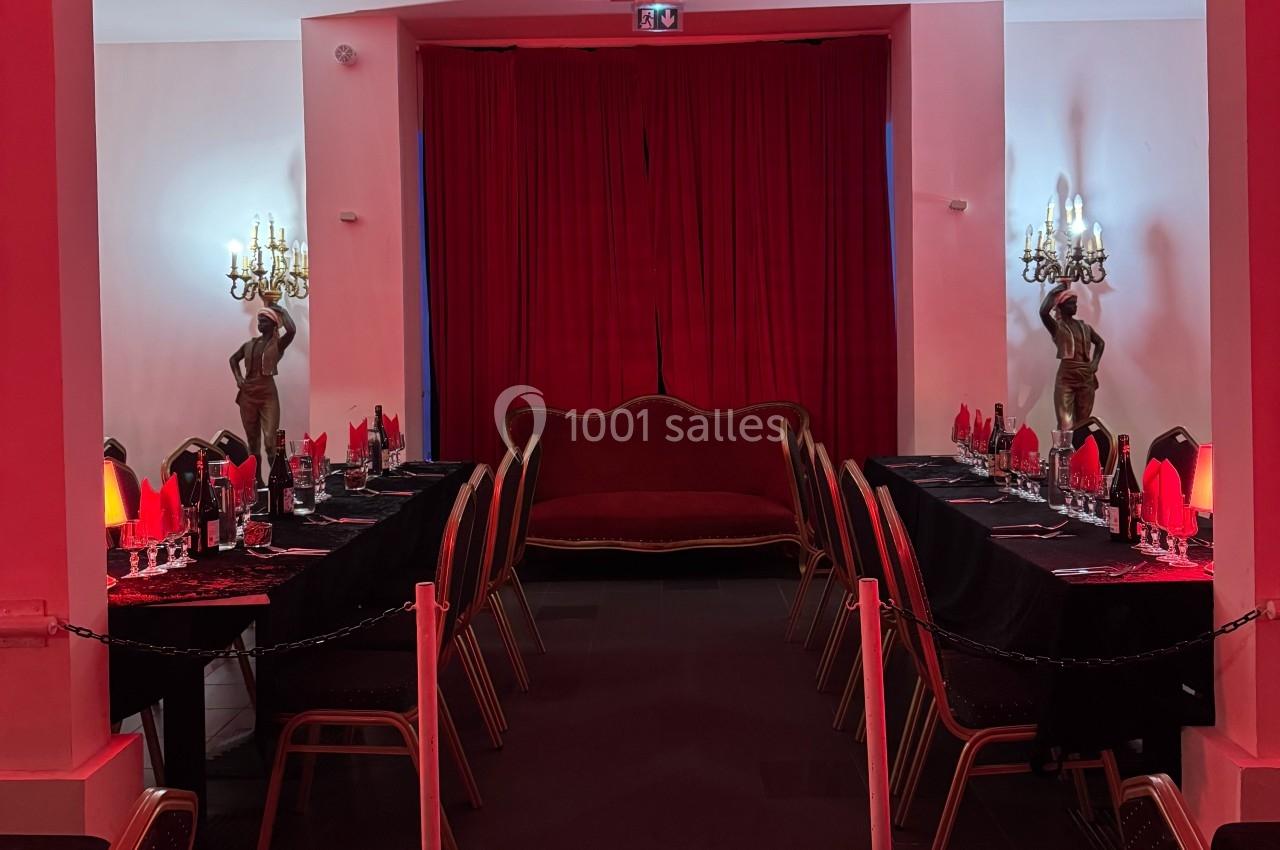 Salle décorée en rouge avec deux longues tables dressées, un canapé central et des statues encadrant des rideaux.