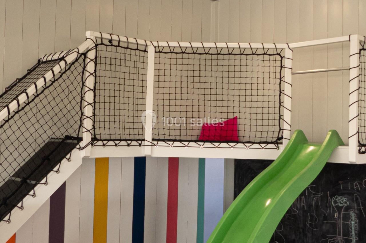 Espace de jeu intérieur avec toboggan vert, coussins colorés au sol et murs décorés de rayures multicolores.
