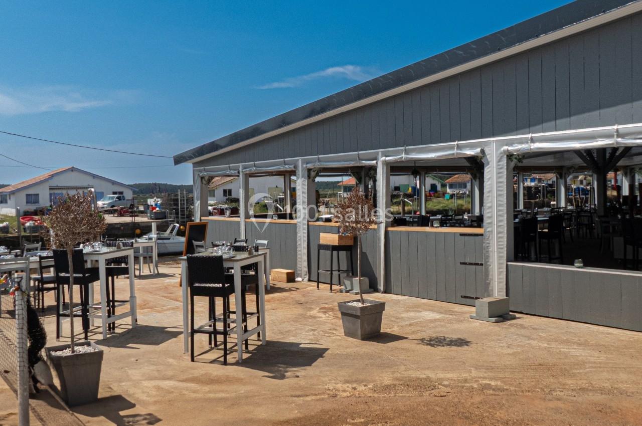 Terrasse extérieure d'un restaurant avec tables hautes, chaises et vue sur des maisons au loin sous un ciel bleu.