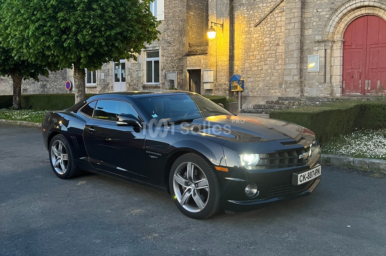Voiture sportive noire garée devant un bâtiment en pierre avec porte rouge, éclairée par une lumière de fin de journée.