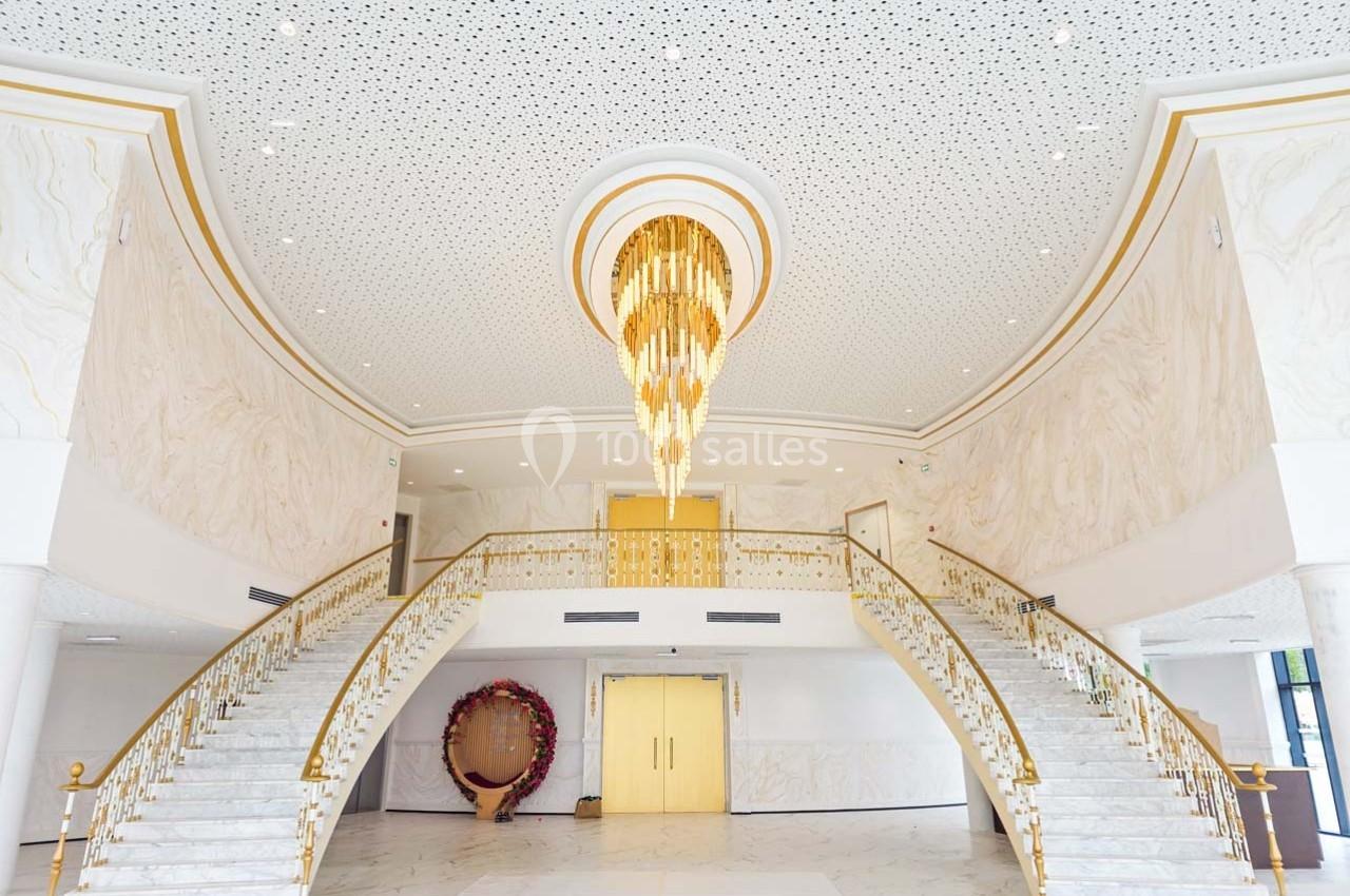 Hall spacieux avec un grand lustre doré, deux escaliers symétriques et des murs décorés de motifs clairs.