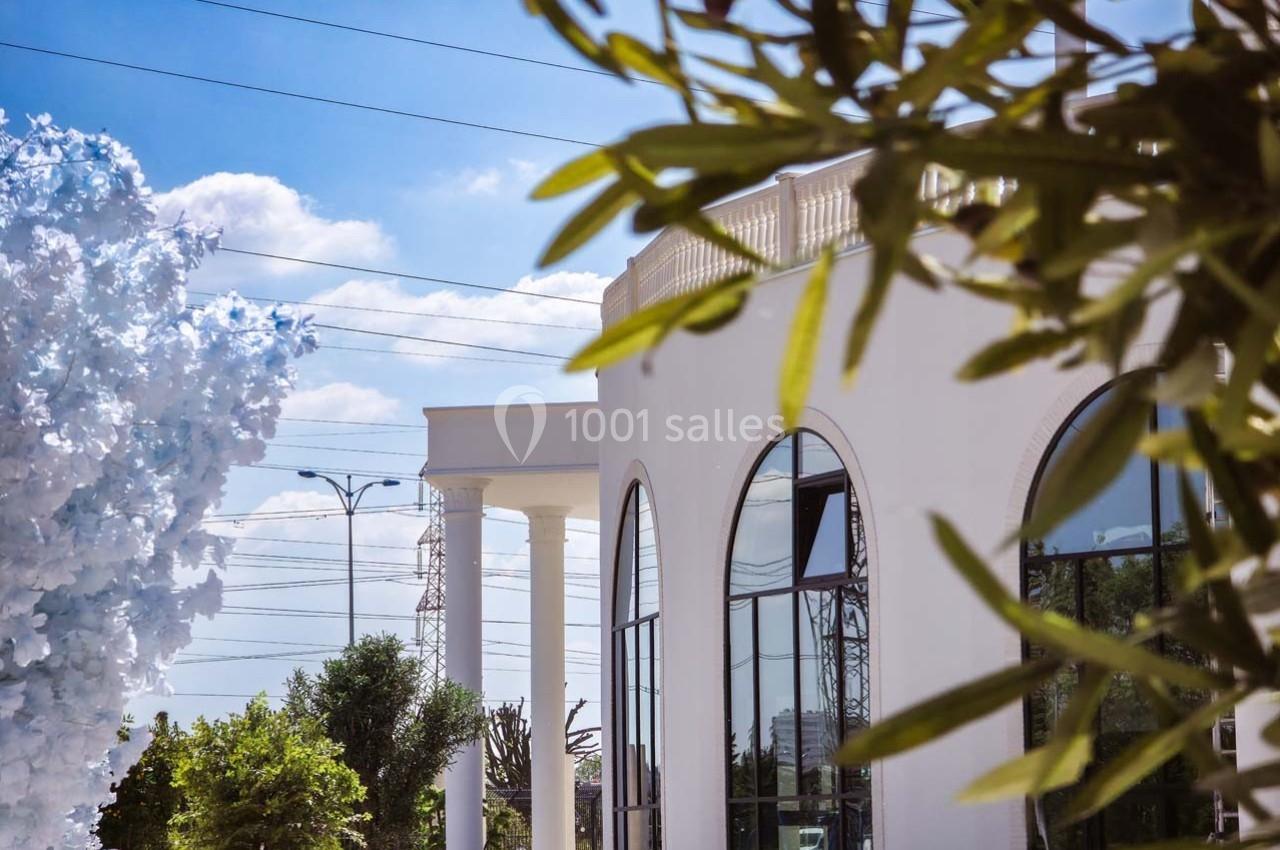 Façade d'un bâtiment blanc avec de grandes fenêtres cintrées, entouré d'arbres et d'un ciel dégagé.