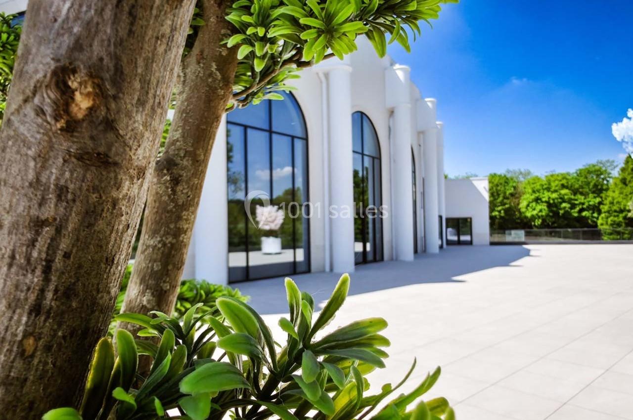 Façade blanche moderne avec grandes baies vitrées, entourée de végétation et éclairée par un ciel bleu dégagé.