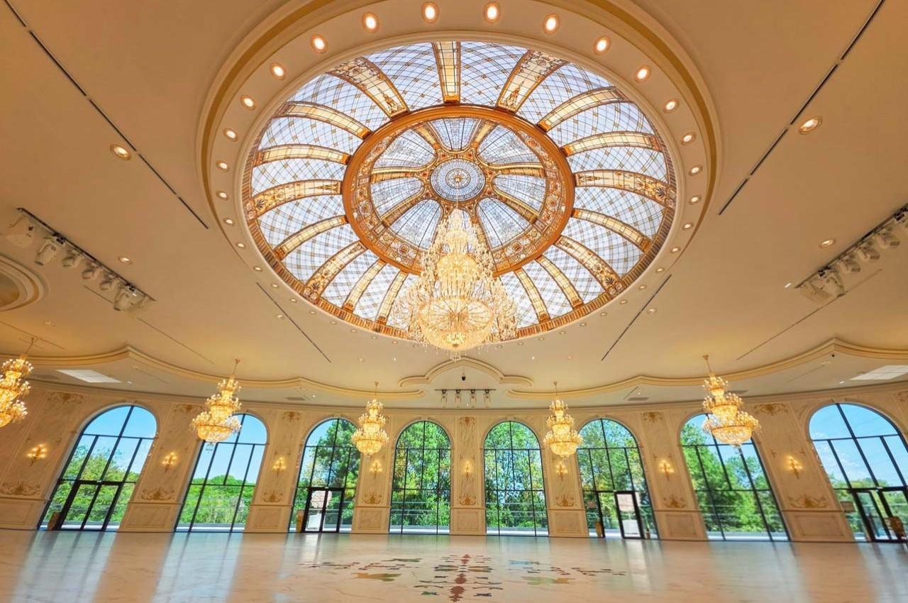 Salle spacieuse avec un plafond en dôme vitré orné de lustres, grandes fenêtres donnant sur un paysage verdoyant.