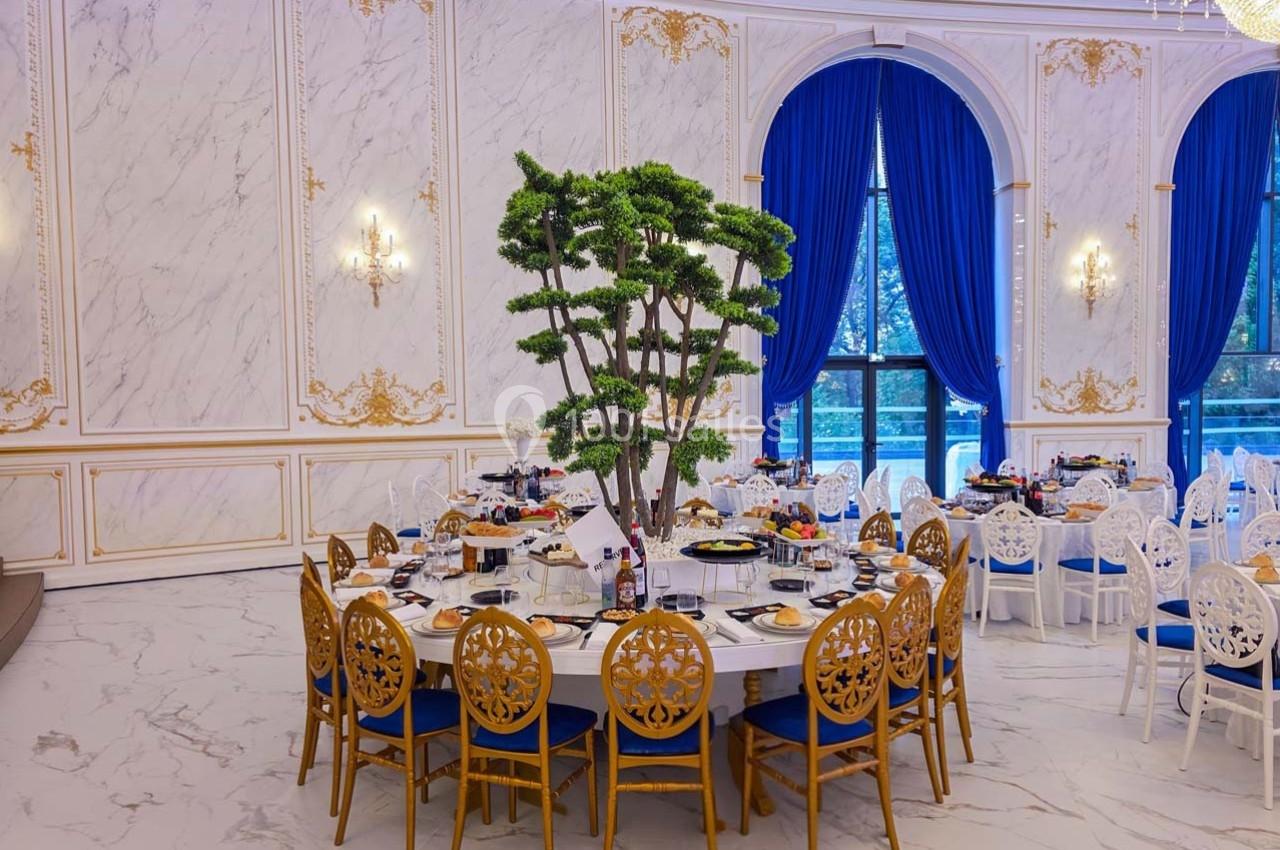 Salle de réception élégante avec tables rondes décorées, chaises dorées et bleues, et un arbre central sur une table.