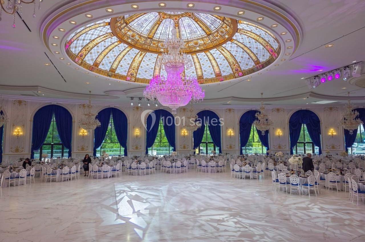 Salle de réception élégante avec un plafond en coupole vitrée, lustres suspendus et tables décorées de nappes blanches.