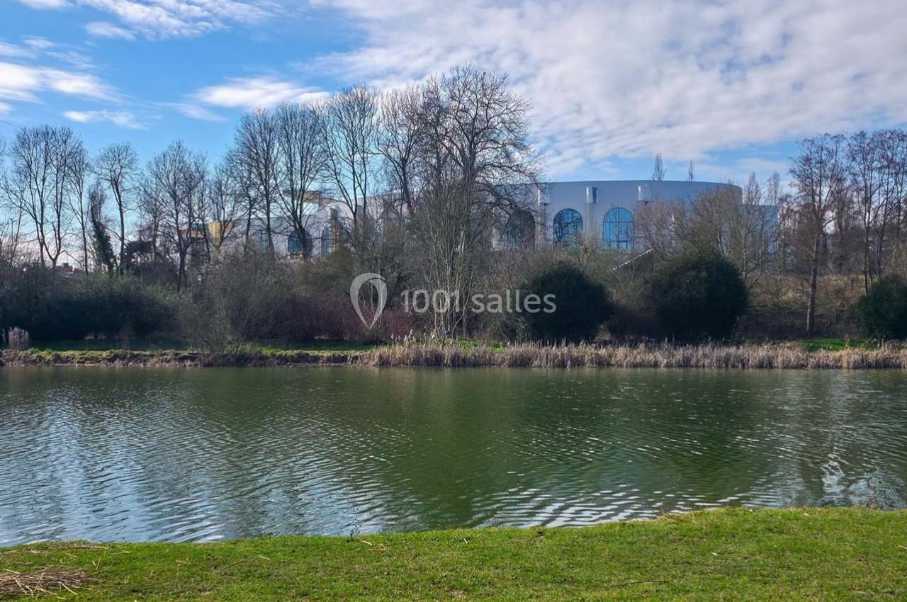 Plan d'eau bordé d'herbe et d'arbres, avec un bâtiment blanc aux grandes fenêtres visible à travers les branches.