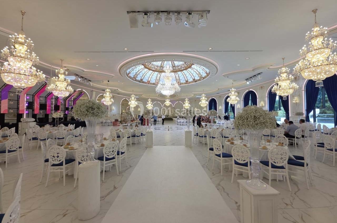 Salle de réception élégante avec lustres imposants, plafond vitré, tables décorées et chaises blanches.