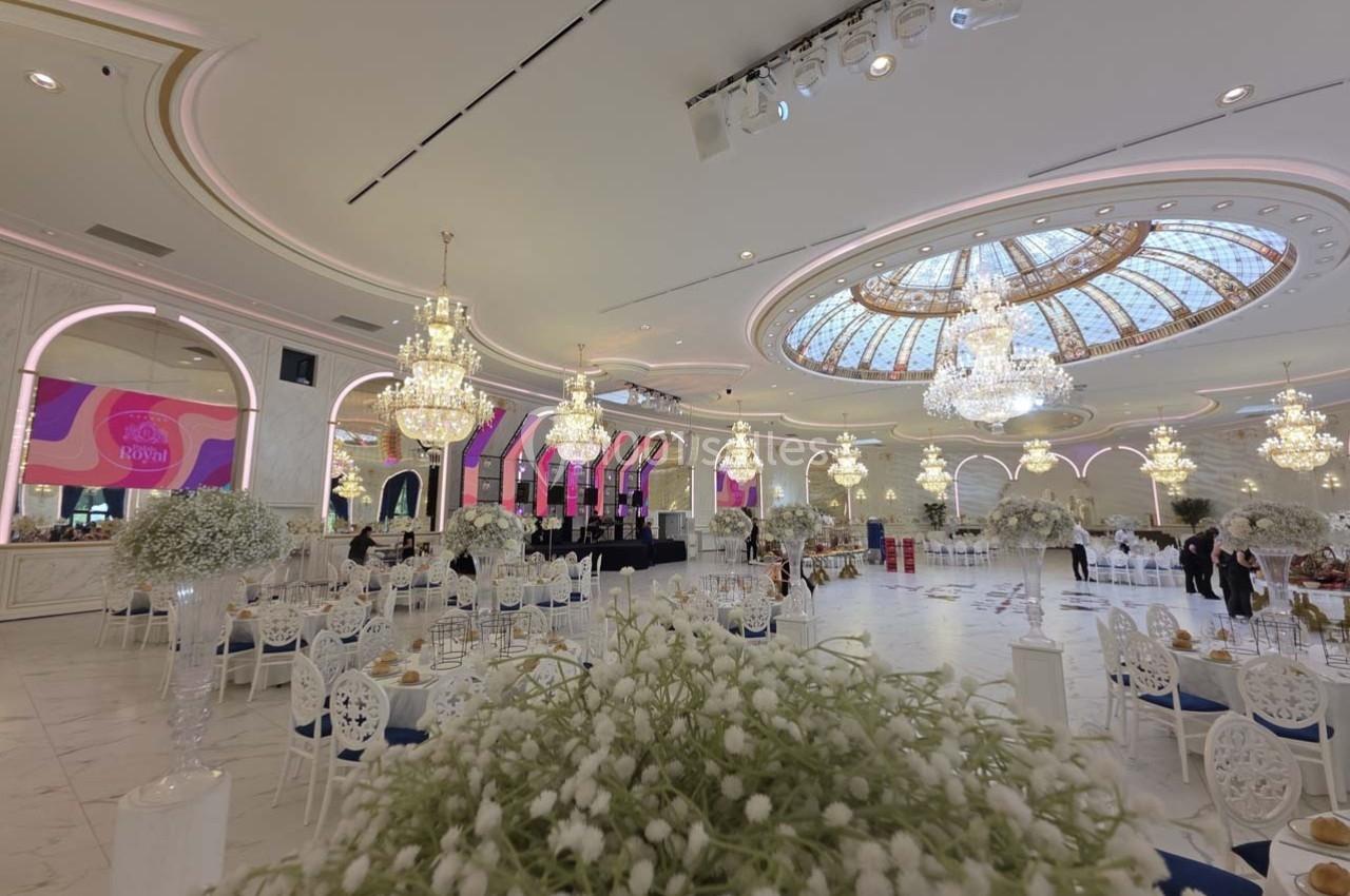 Salle de réception élégante avec lustres, verrière, tables décorées de fleurs blanches et scène en arrière-plan.