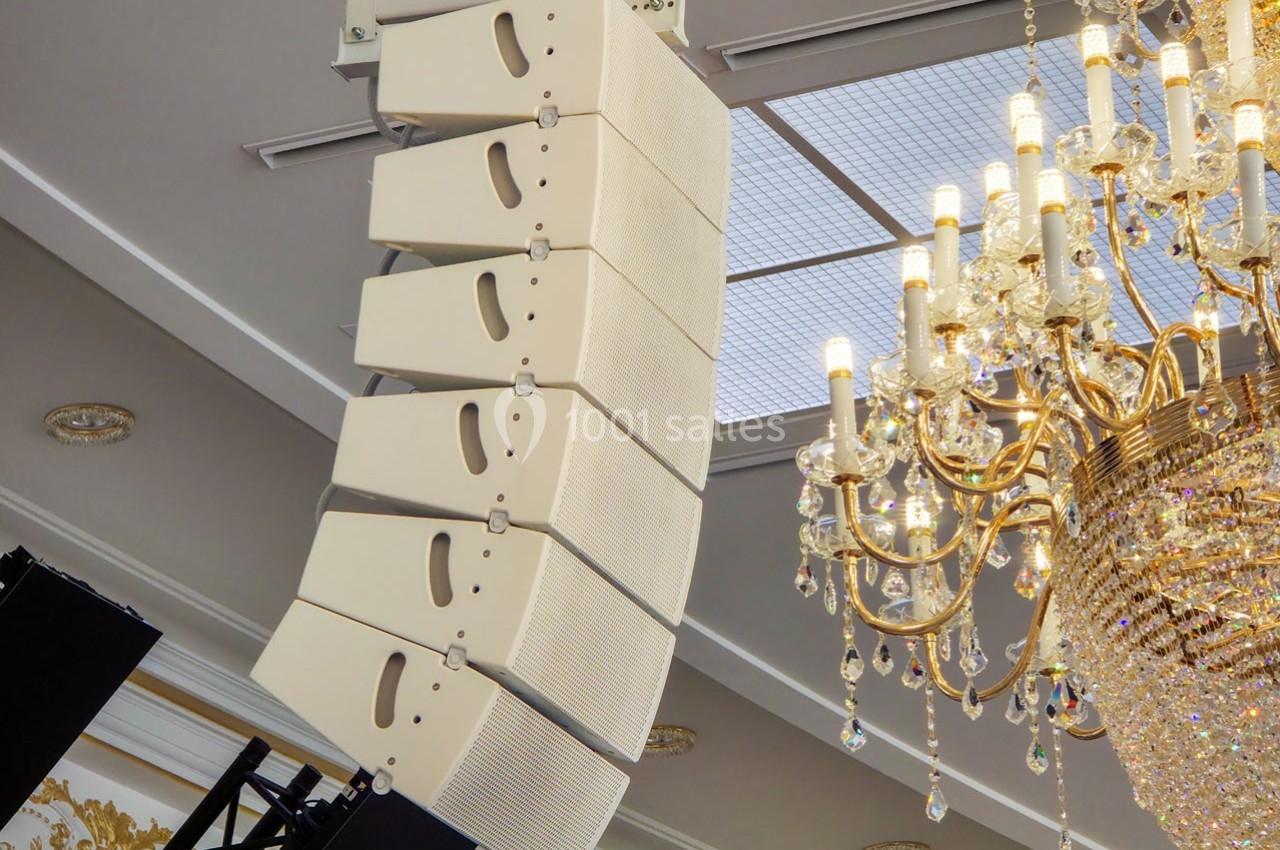 Enceintes suspendues en ligne array près d'un lustre dans une salle élégante avec plafond lumineux.