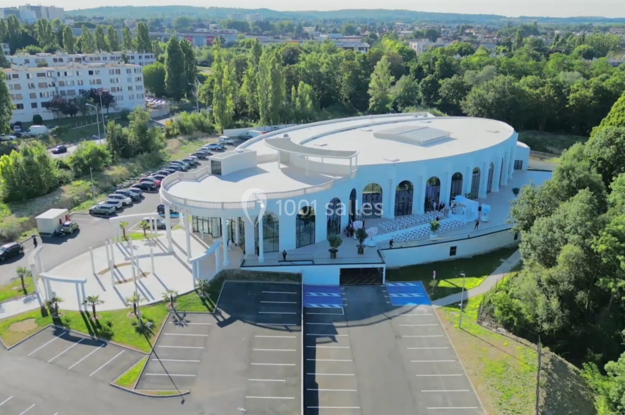 Bâtiment blanc moderne de forme ovale avec de grandes baies vitrées, entouré de végétation et de parkings.