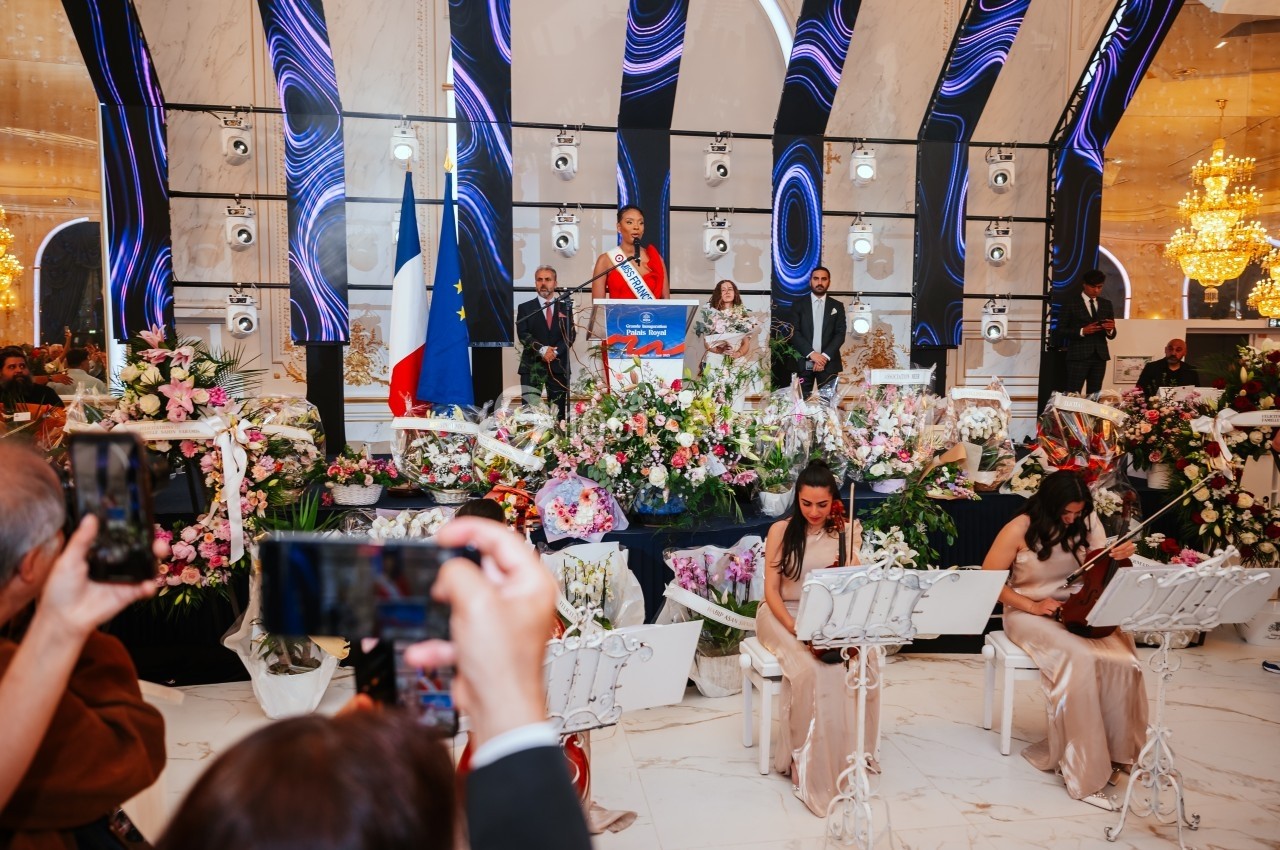 Une femme s'exprimant sur une scène décorée, entourée de musiciens et de fleurs, avec des drapeaux en arrière-plan.