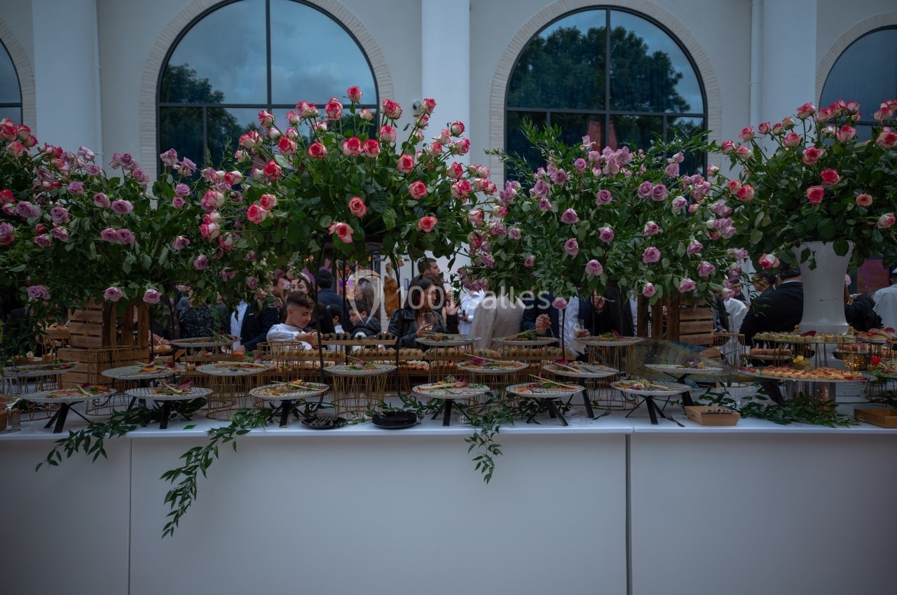 Buffet décoré de fleurs roses avec des plats variés, dans une salle lumineuse avec des fenêtres en arc.