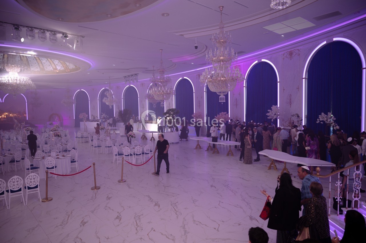 Salle de réception élégante avec des invités, des tables décorées, des lustres et un éclairage violet tamisé.
