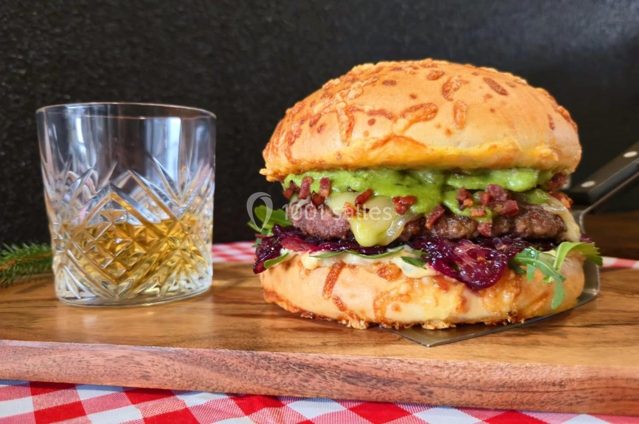 Burger garni de viande, fromage, avocat et sauce, posé sur une planche en bois avec un verre de whisky à côté.