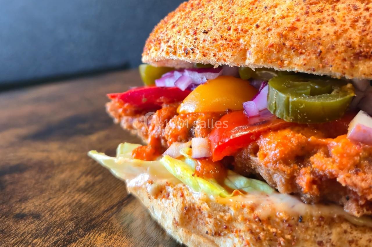 Sandwich garni de poulet frit, légumes colorés, jalapeños et sauce, présenté sur une table en bois.