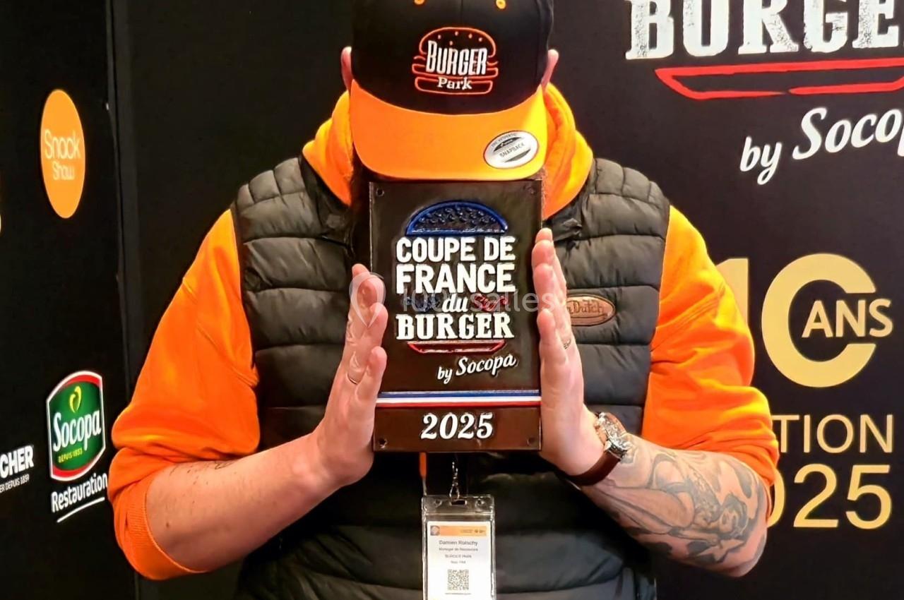 Un homme tenant un trophée marqué ’Coupe de France du Burger 2025’ devant son visage, en tenue orange et noire.