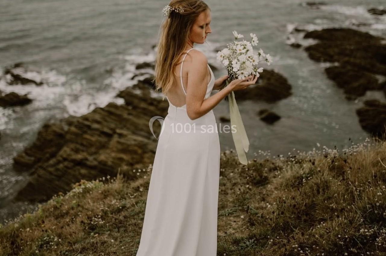 Une femme en robe blanche tient un bouquet de fleurs, debout sur une falaise surplombant la mer.