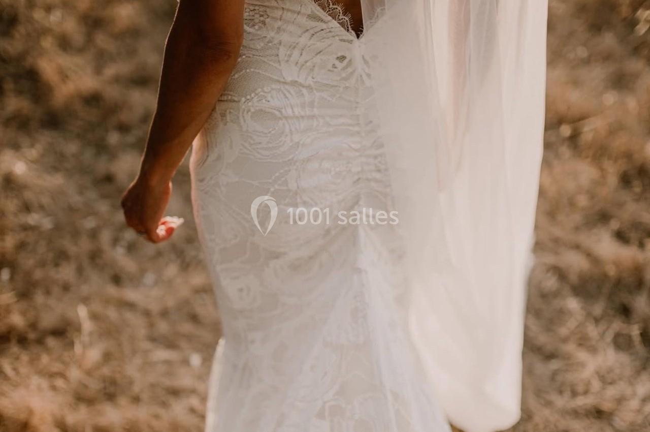 Femme de dos portant une robe de mariée en dentelle et un voile, debout sur un sol herbeux et sec.