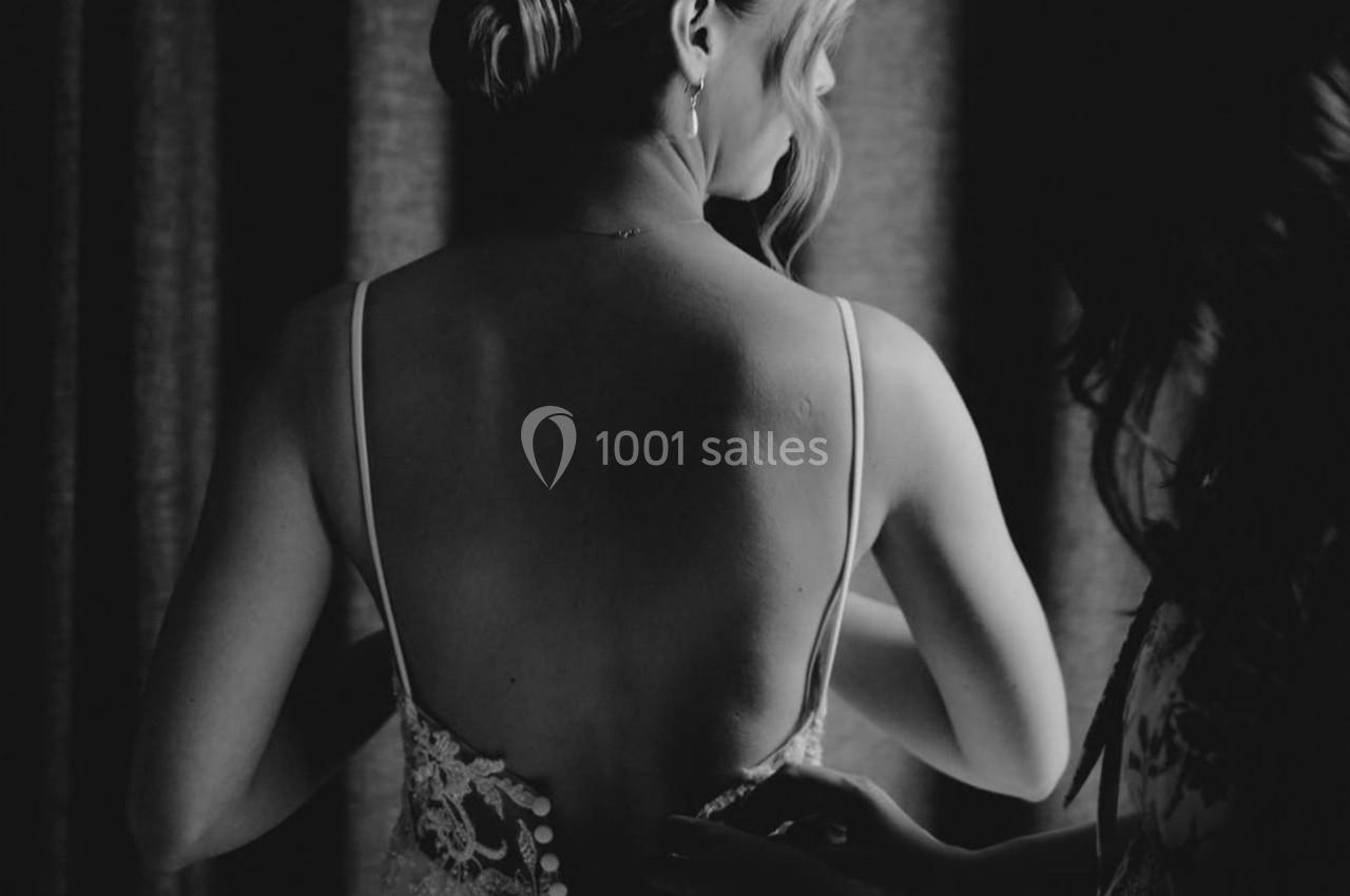 Une femme en robe de mariée est vue de dos, tandis qu'une autre personne ajuste les boutons de sa tenue.