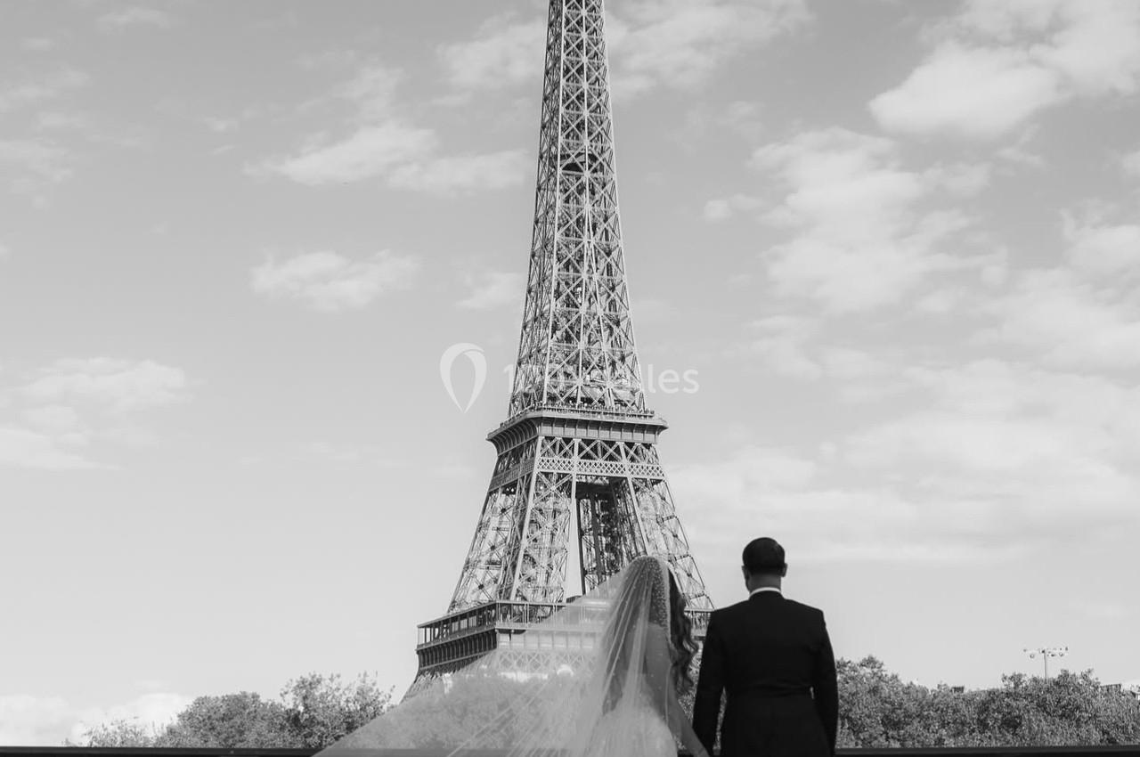 Un couple en tenue de mariage regarde la tour Eiffel depuis un pont par une journée ensoleillée.