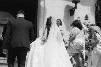 Un couple monte des marches, la mariée en robe blanche et voile, entourée d'invités élégamment vêtus.