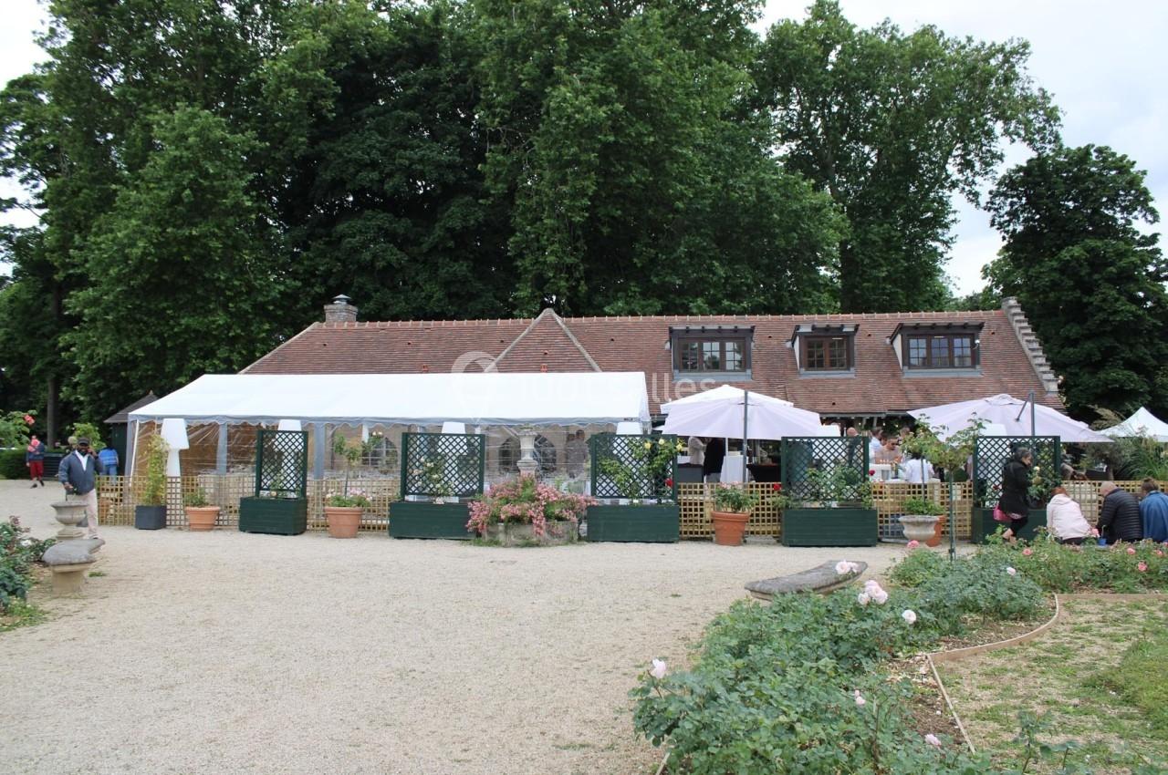 Grande maison en pierre avec toit en tuiles, entourée de tentes blanches, parasols et végétation luxuriante.