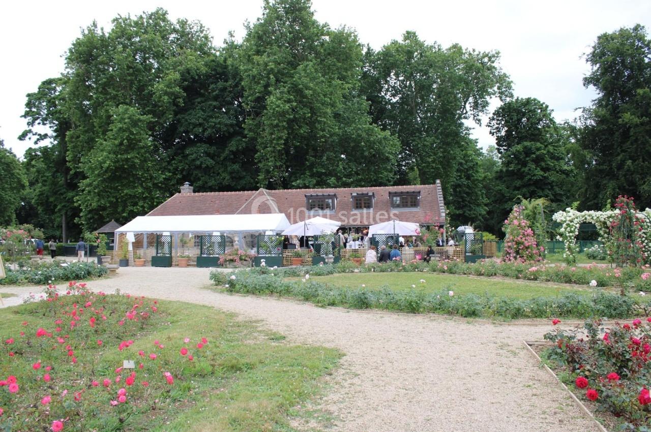 Maison entourée de rosiers et de grands arbres, avec une terrasse couverte et des tables installées à l'extérieur.